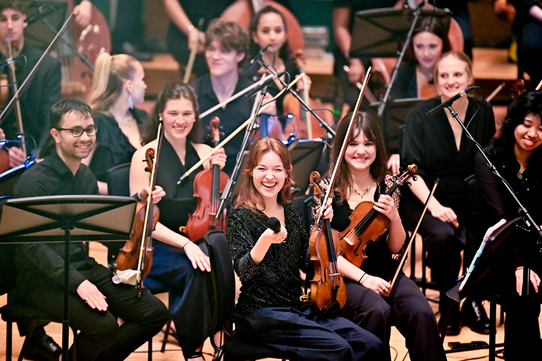 A group of violin students, wearing smart attire and sitting in seats, smiling at the camera.